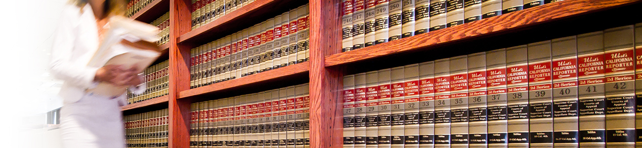 A person in a white lab coat walks through a room filled with books on shelves, suggesting a scholarly environment.
